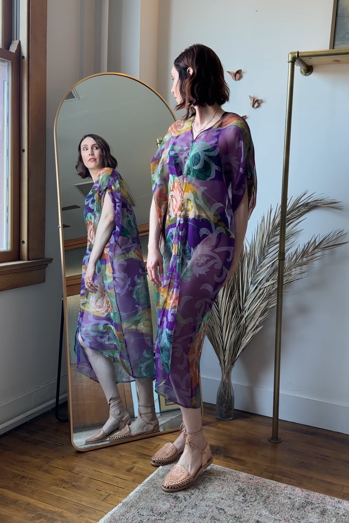 Woman in a colorful dress standing in front of a full-length mirror in a room with a plant and window.