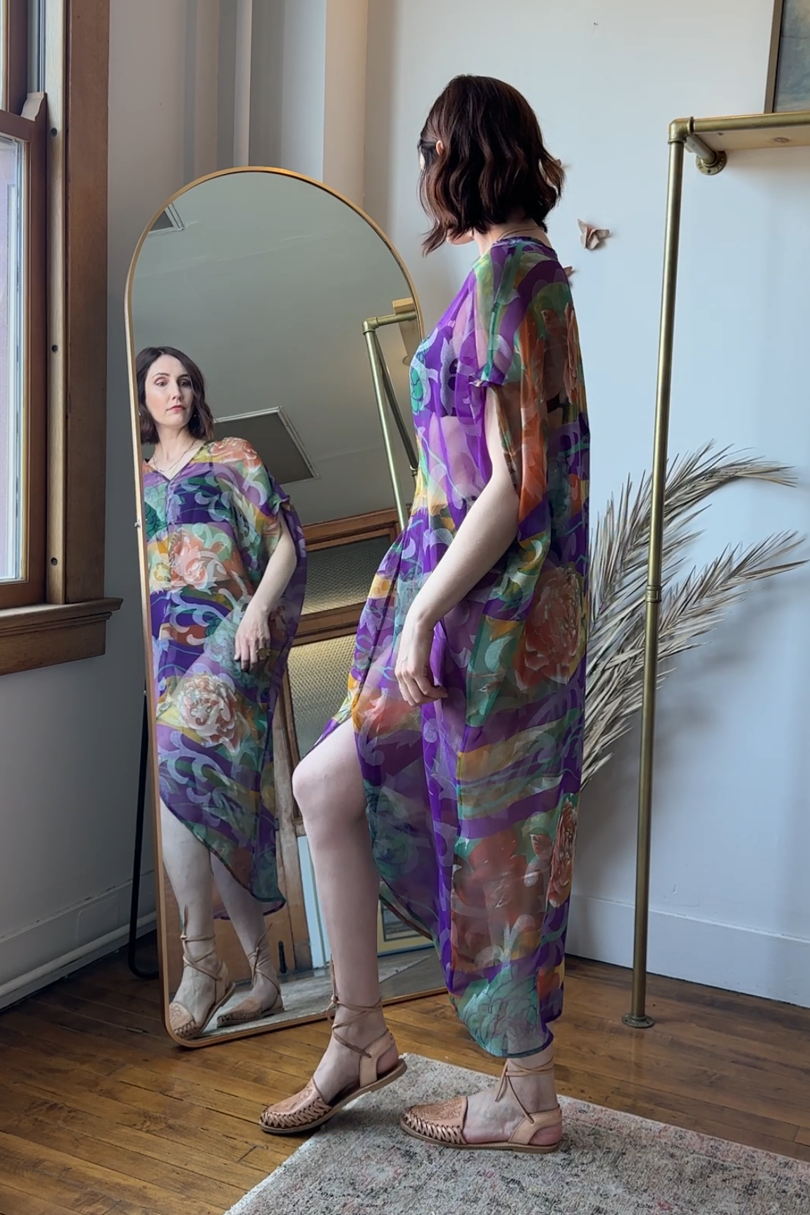 Woman in a colorful dress standing in front of a full-length mirror in a room with wooden flooring and a plant.