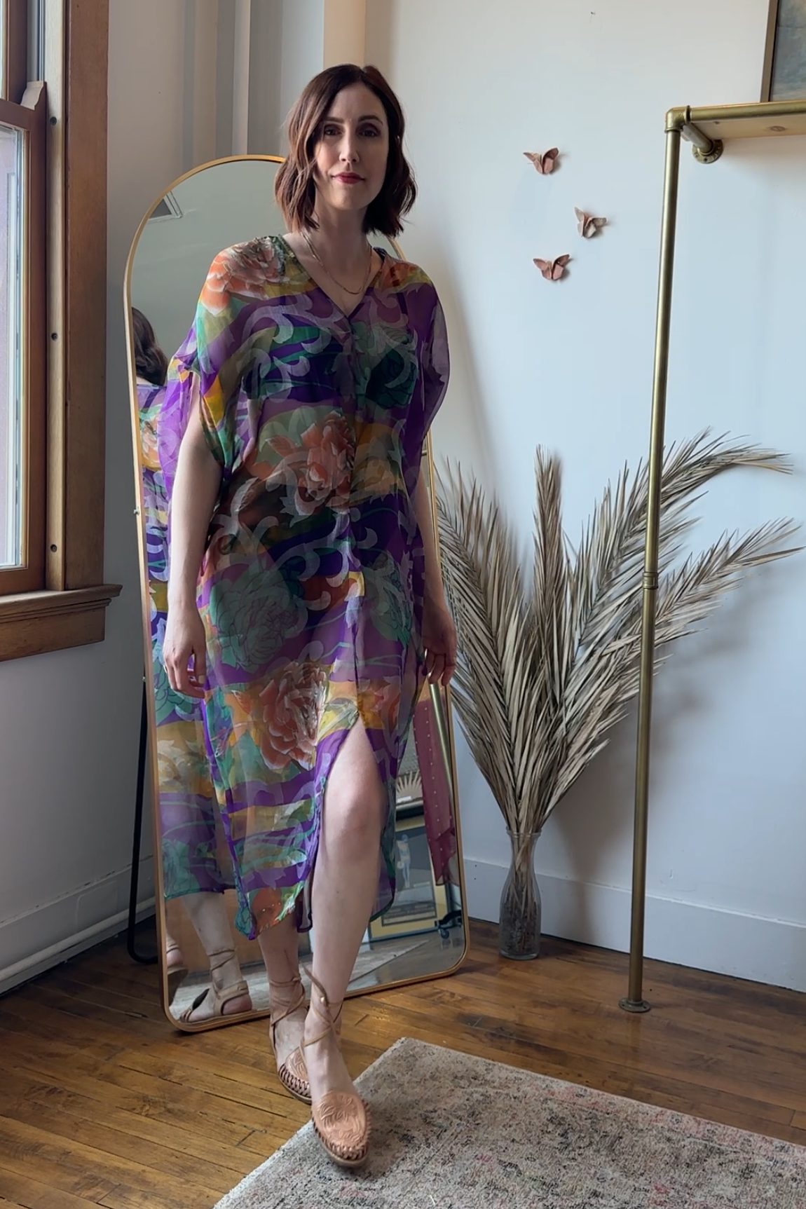 Woman in a colorful dress standing in front of a mirror in a room with a plant and coat rack.